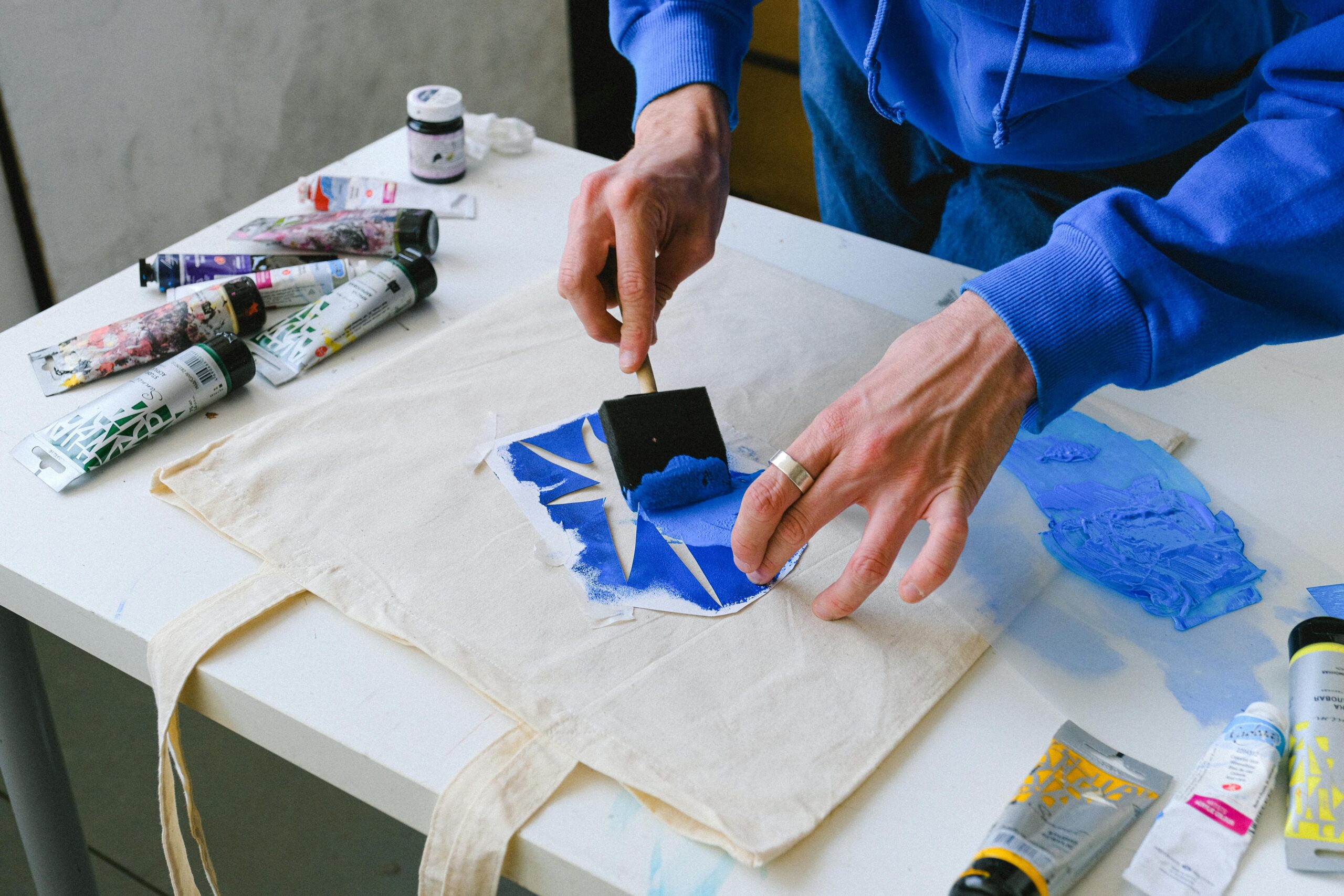 Artist applies blue paint to canvas bag using a stencil technique in a creative studio setting.